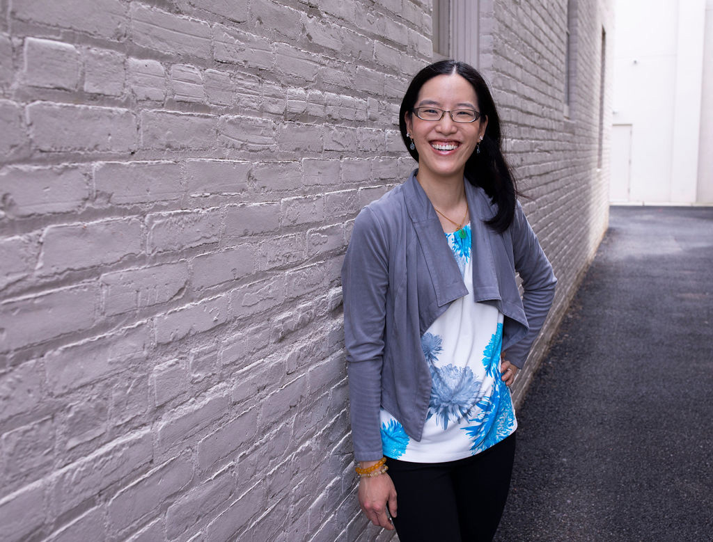 An Asian woman with a grey jacket standing next to a white brick wall.