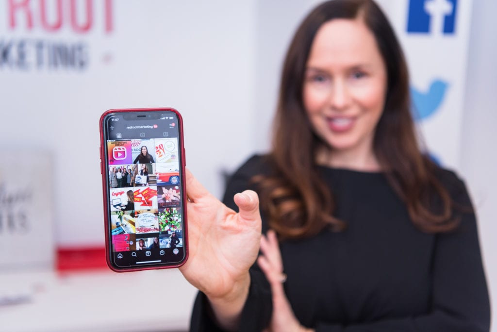 A woman in a black blouse holding a phone showing her Instagram's profile grids.