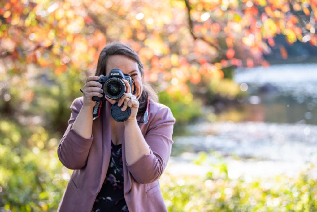 A woman holding a Canon DSLR and taking a picture at a park.