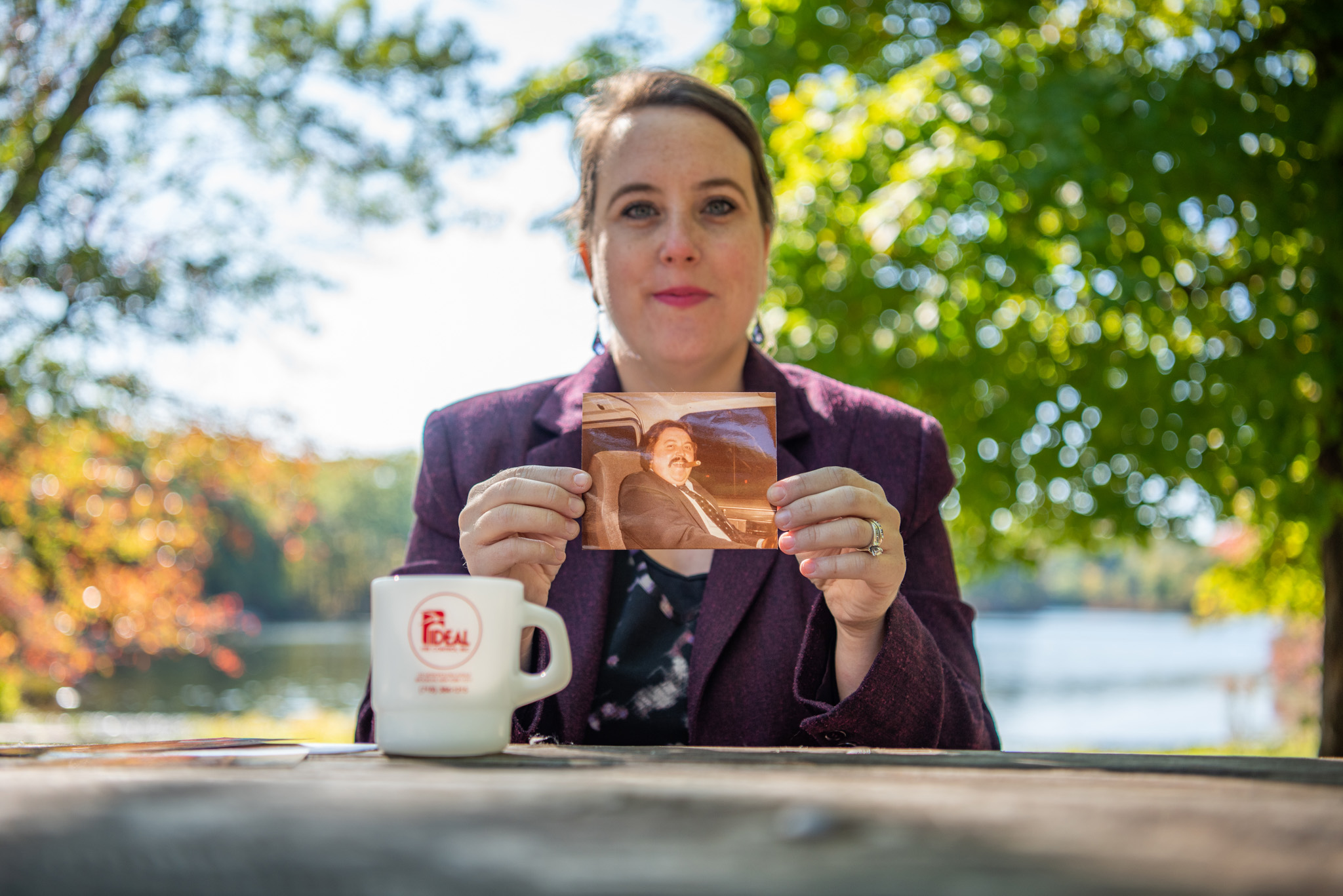 A woman holding a picture of her father sitting at a park picnic table.