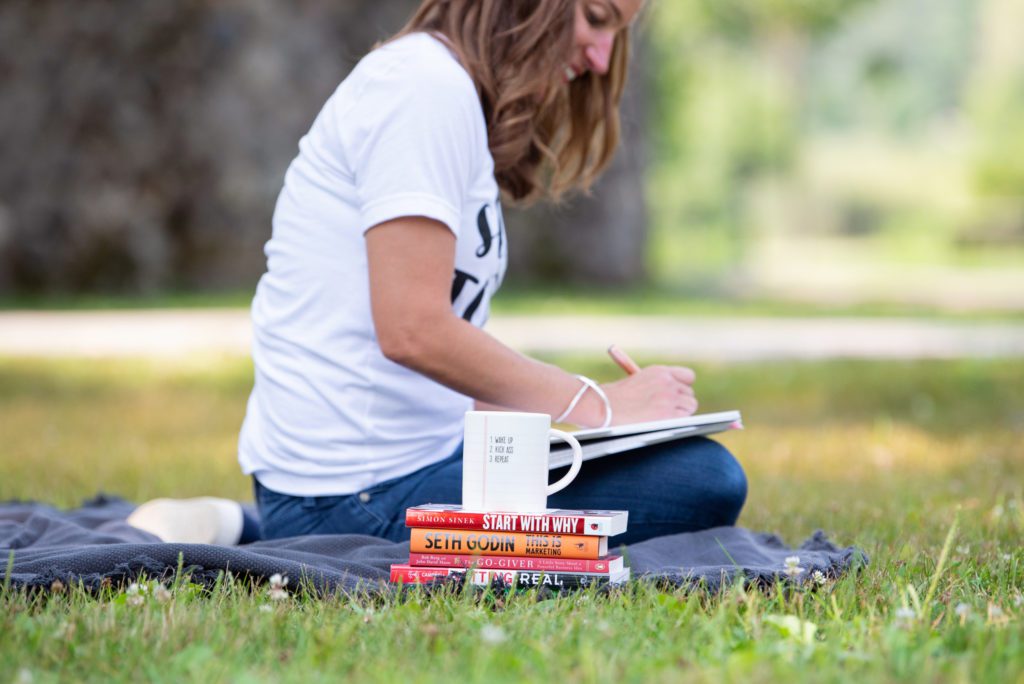 A Caucasian woman is at a park, sitting on a blanket writing with her books and mug on the side.