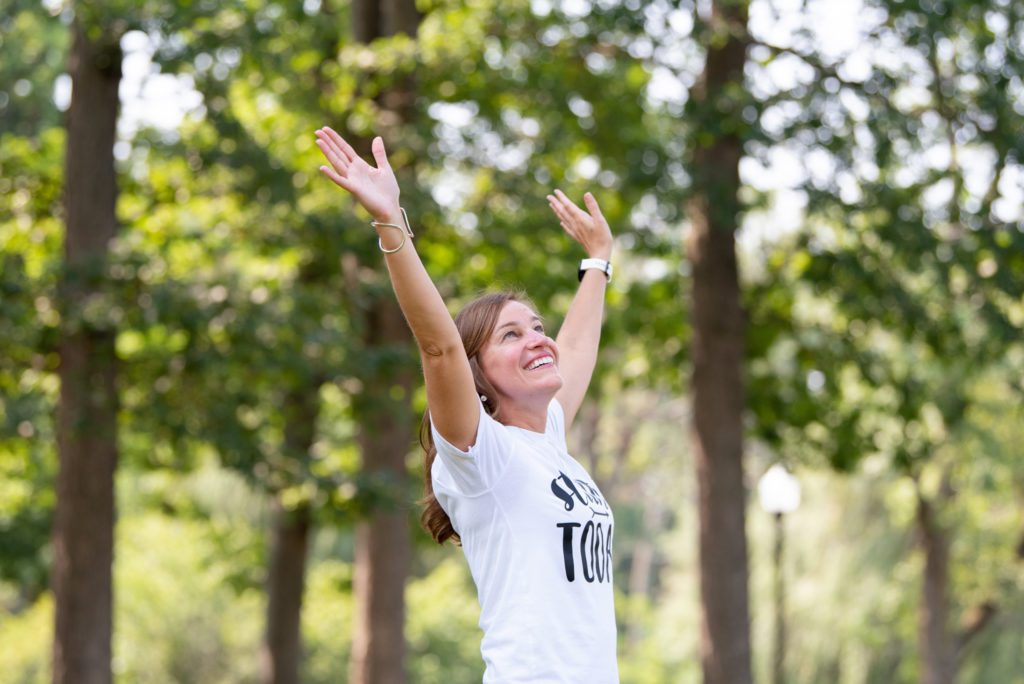 A Caucasian woman is happy and putting both of her arms up at a park.