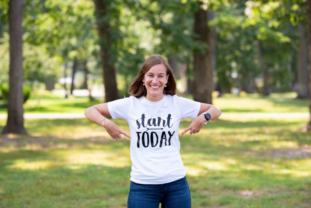 A Caucasian woman is pointing with both of her hands on her "Start Today" T-shirt.