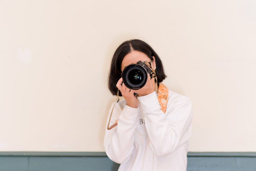 A woman holding a DSLR camera with a white sweatshirt.