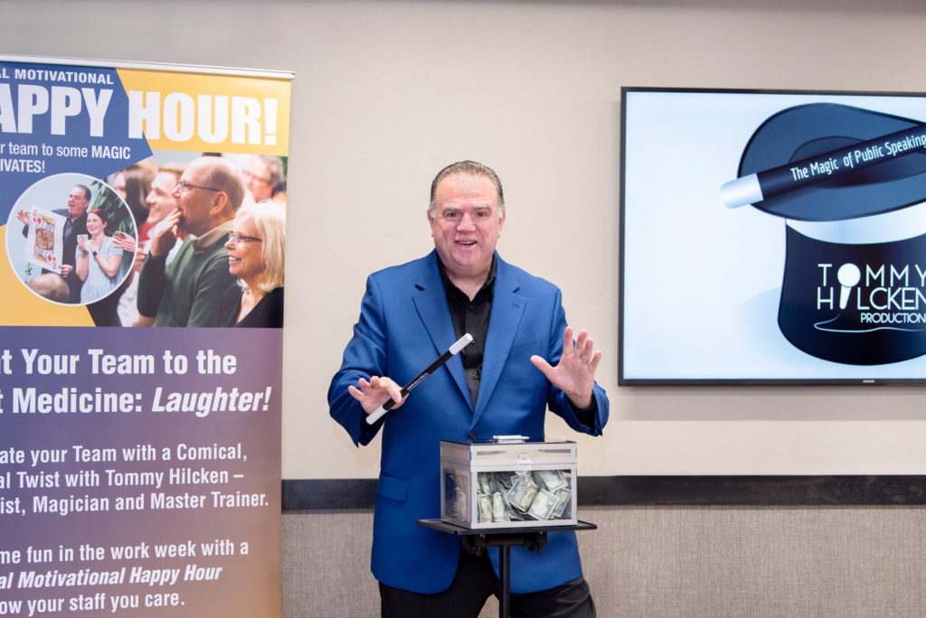 A man in a blue jacket holding a magic wand and a box of cash doing magic trick.