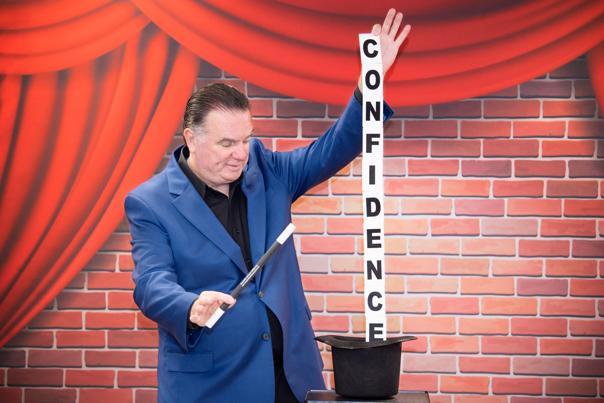 A man in a blue jacket holding a magic wand and pulling out the word "Confidence" out of the magic hat.