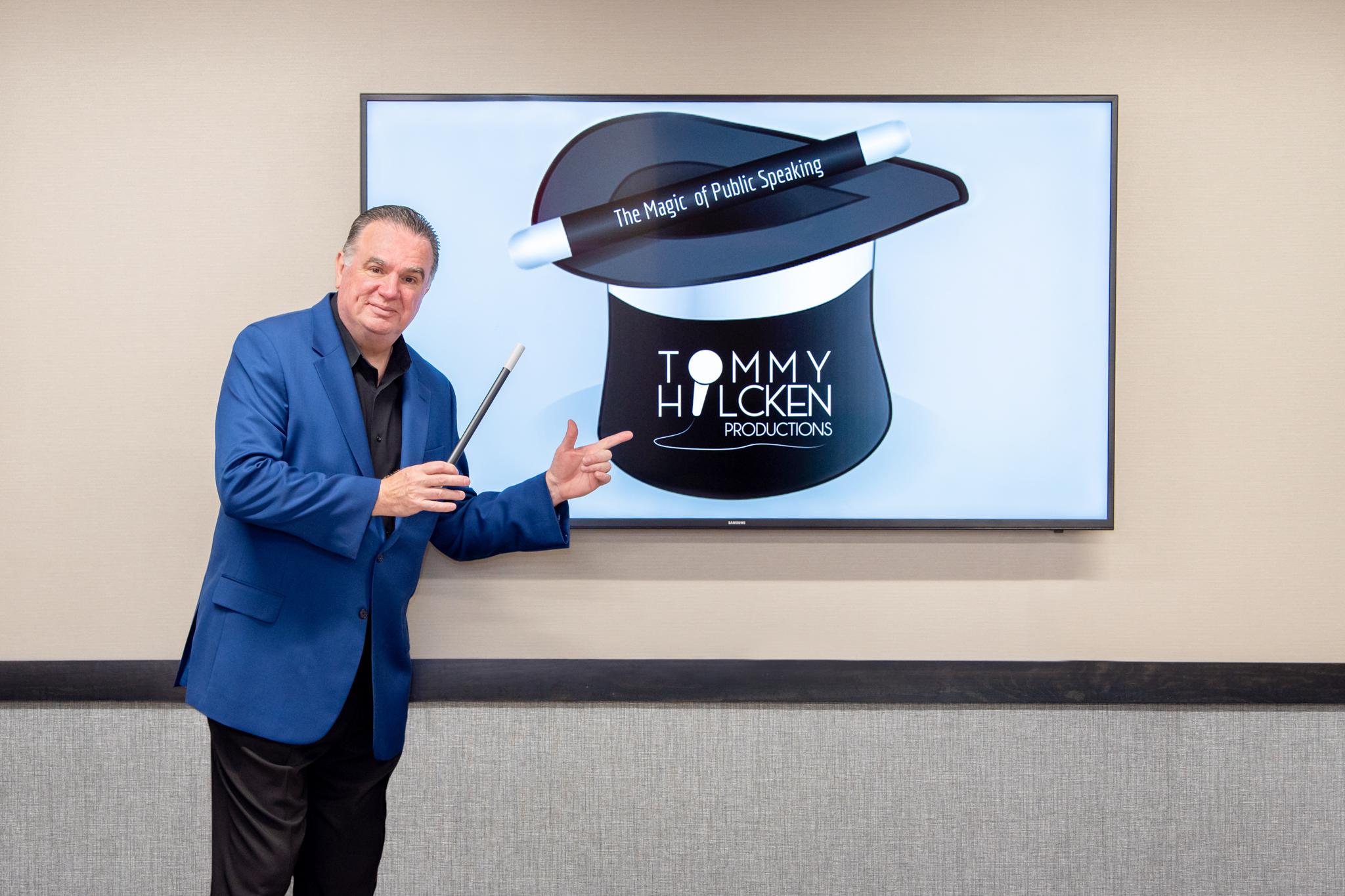 A man in a blue jacket holding a magic wand and pointing to the TV showing Tommy Hilcken Productions logo.