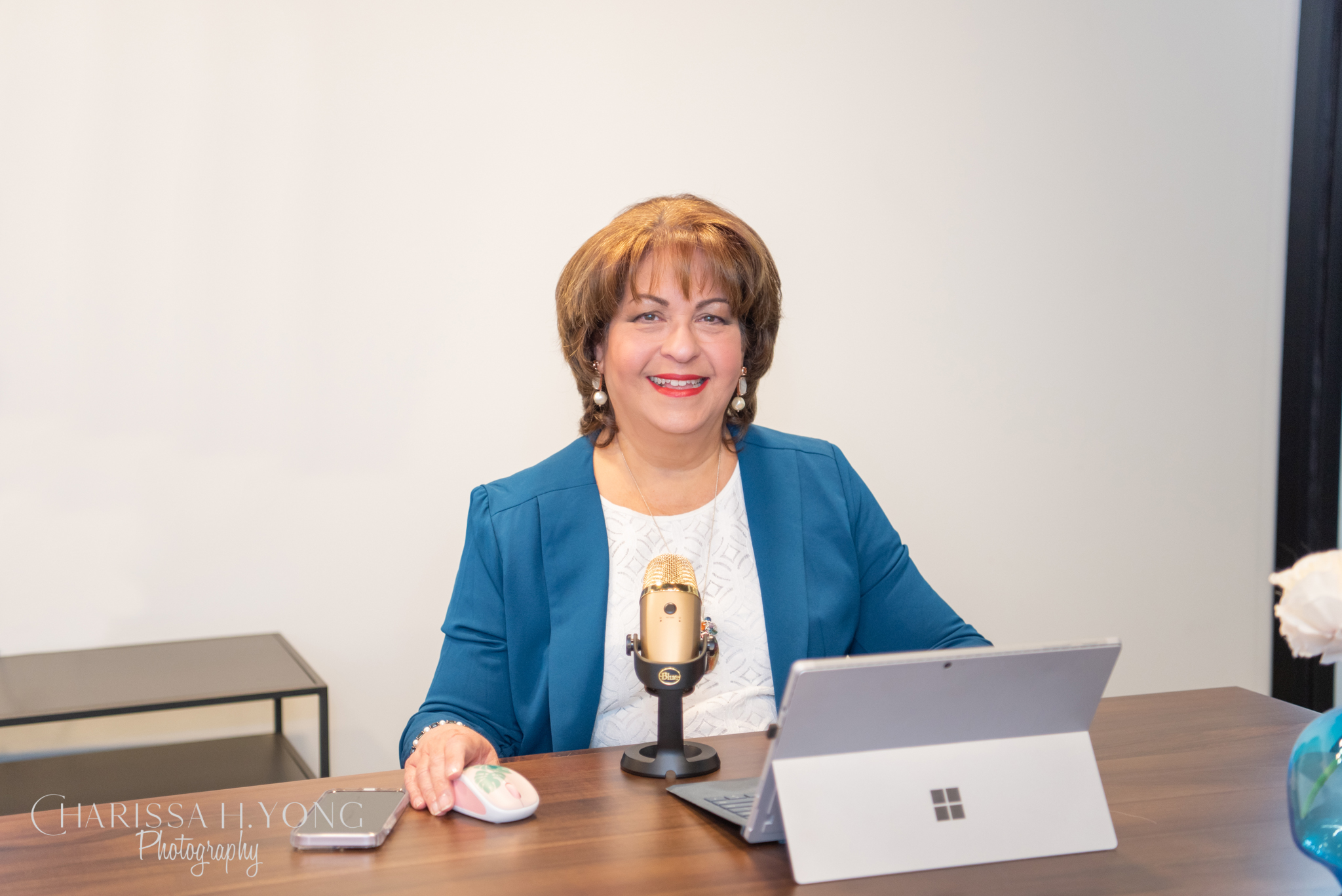 A middle age woman in the blue jacket sitting on the desk working with her gold microphone.