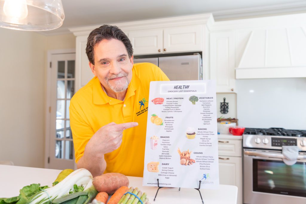A male health coach pointing to a healthy groceries list info board in his kitchen.