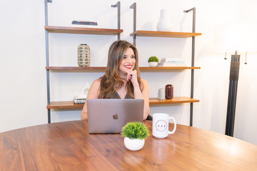 A woman sitting on a desk with her laptop and mug thinking about hiring a branding photographer.