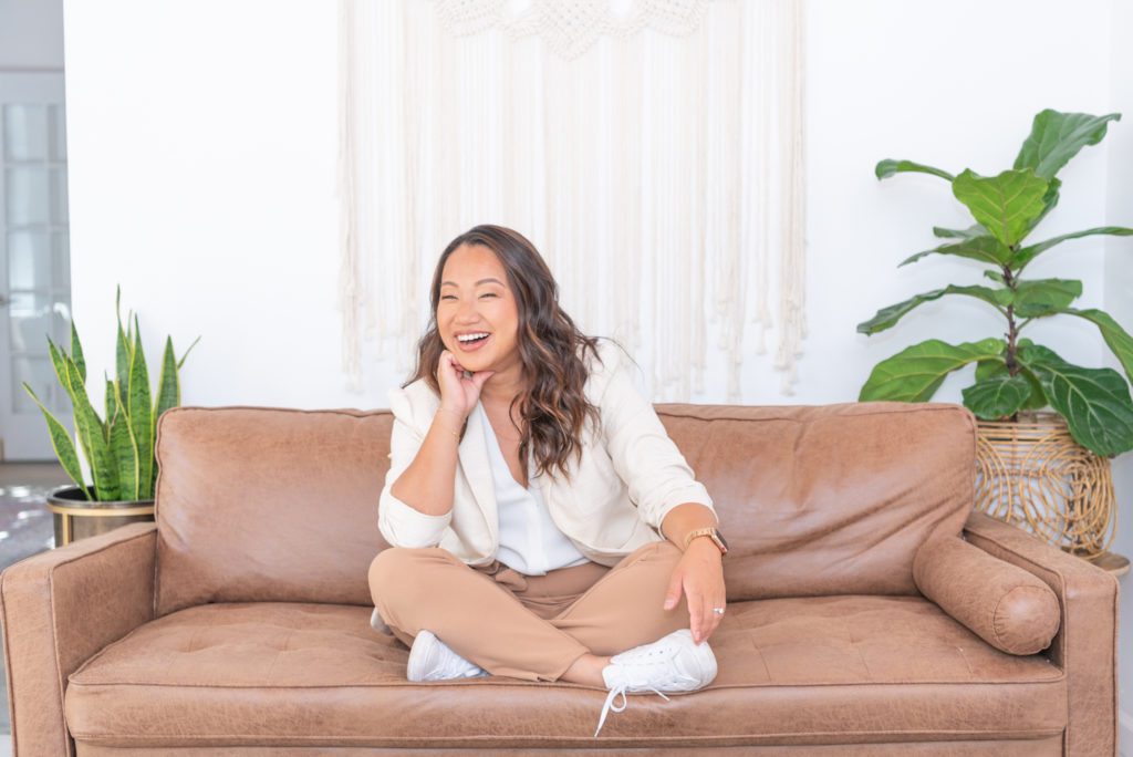 An Asian female is sitting criss cross on a couch and smiling.