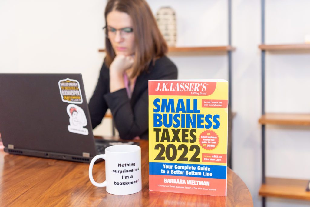 A female bookkeeper sitting and working on her laptop with a Small Business Taxes 2022 and a mug in front of her laptop.