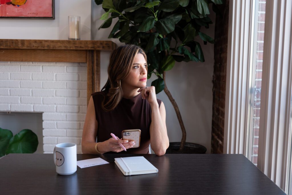 A Latino CEO woman in a brown dress holding her phone in her office, notebook, pen, notepad, and a mug on her desk, and she's deep in thoughts looking out the window.