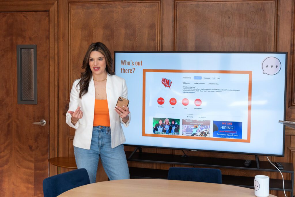 A Latino CEO woman standing and smiling while presenting about her marketing agency at a co-working space.