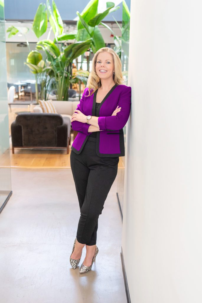 A female woman business owner with a purple blazer leaning against the wall with her arms crossed and a friendly smile posing for her branding photoshoot.
