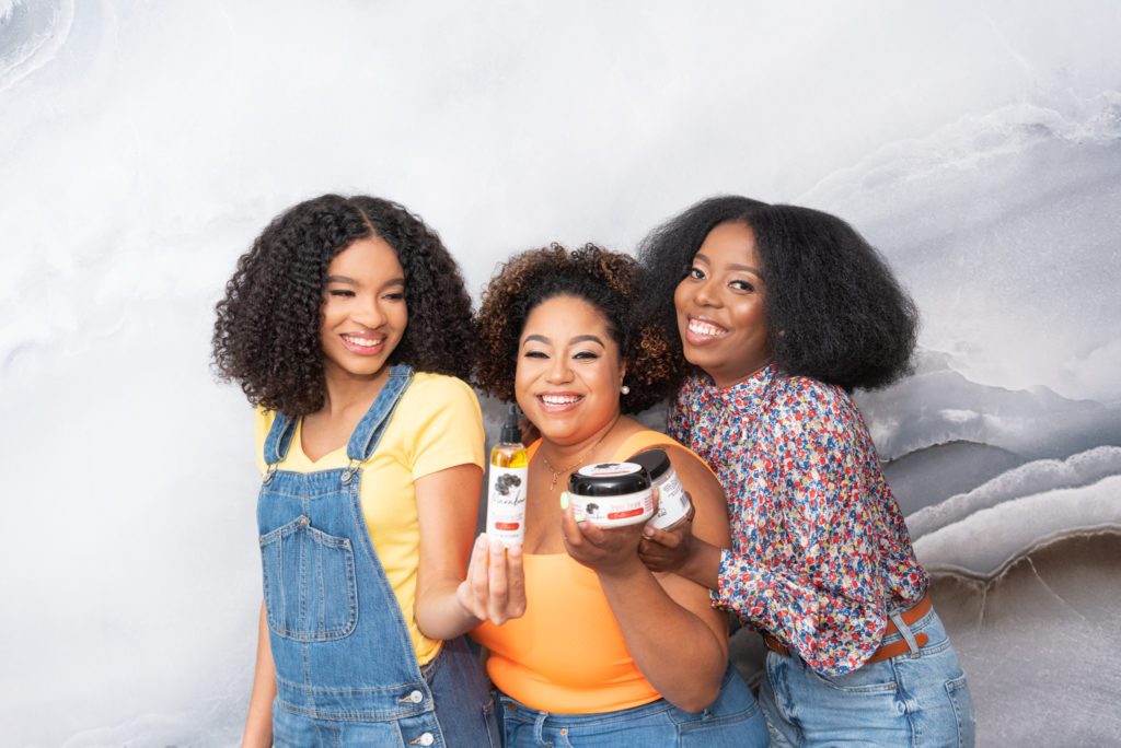 A group of African American women holding a line of natural hair care products and laughing.