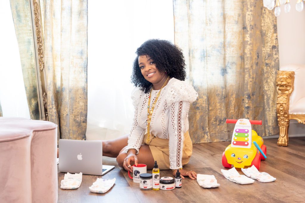 An African American momtrepreneur sitting on the floor with her natural hair care products, laptop, mug, diapers and baby toy.