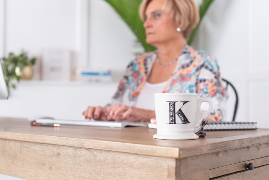 A female health and wellness coach typing on her computer with a "K" letter mug in the foreground.