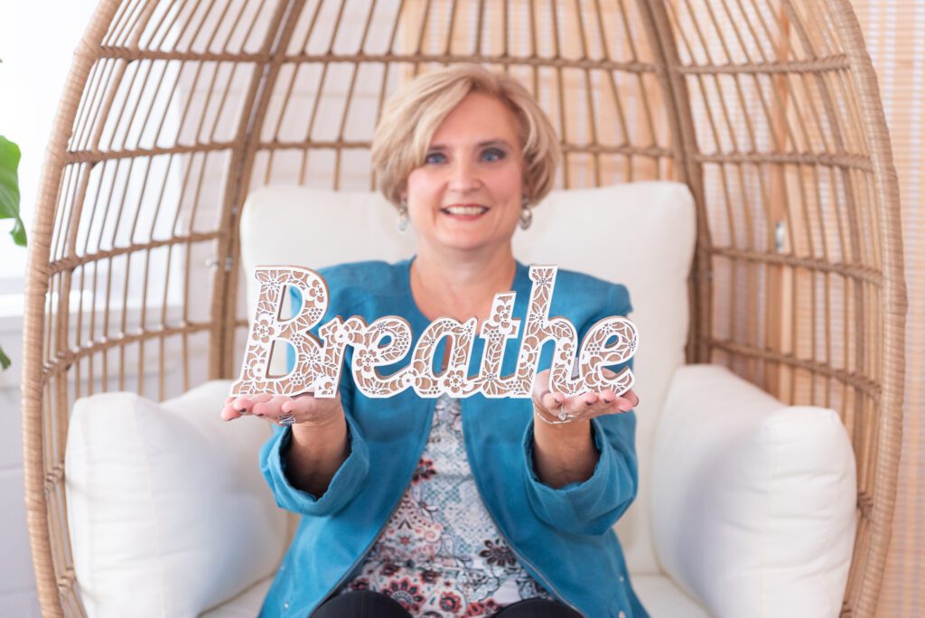 A female health and wellness coach holding a "Breathe" sign and sitting in an egg chair.