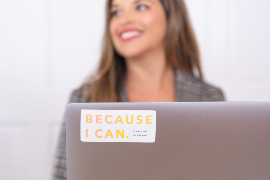 A picture of a sticker on a laptop with a woman in the background with the words "Because I can."