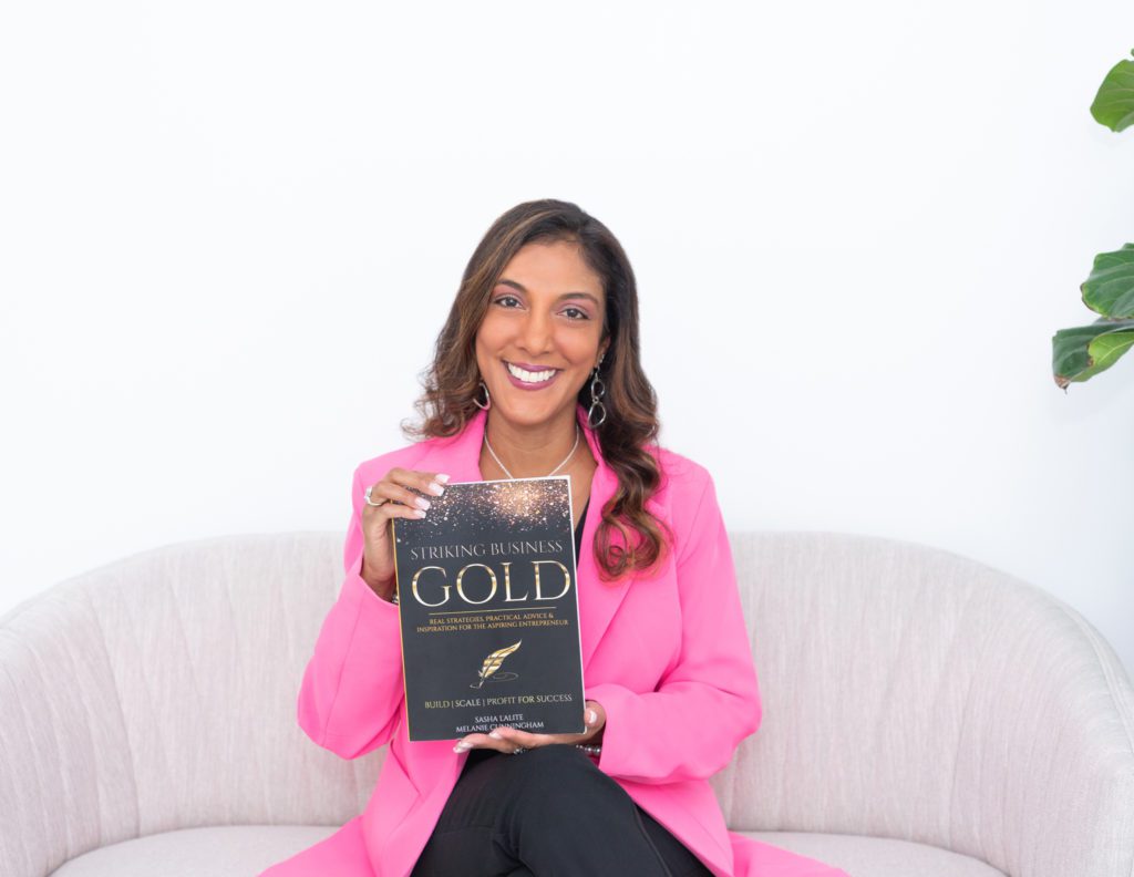 An African American Woman with a pink jacket sitting and holding a book she co-wrote called Striking Business Gold.