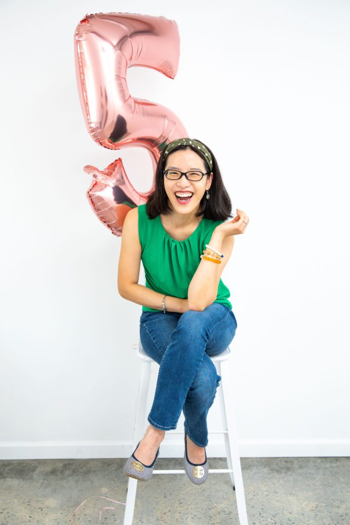 An Asian female sitting on a stool with a number 5 rose gold balloon behind her.