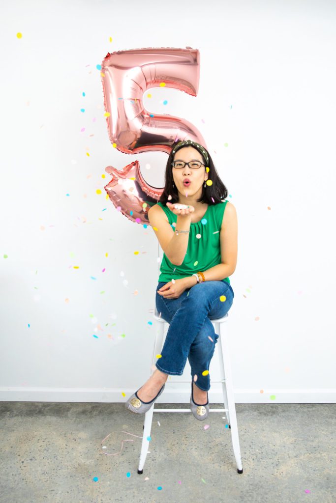 An Asian female sitting on a stool with a number 5 rose gold balloon behind her and she's blowing confetti to celebrate 5 years in business.
