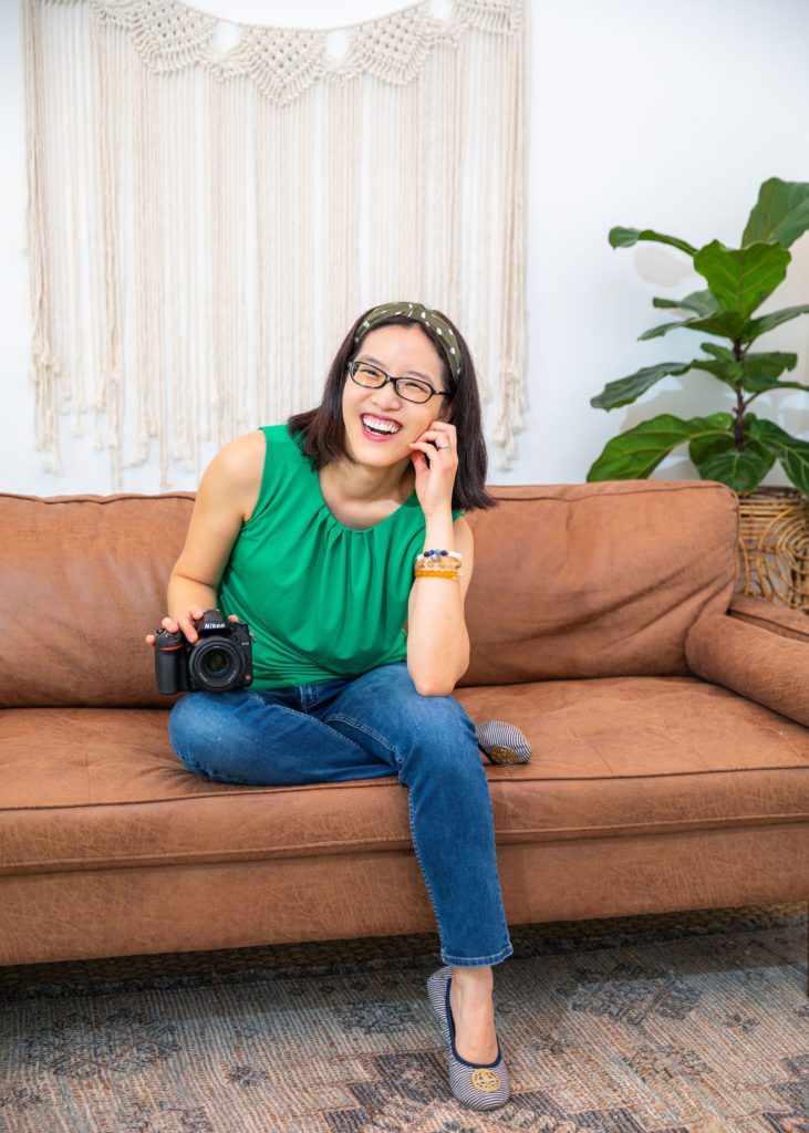 An Asian female personal branding photographer wearing a green top smiling and sitting on the sofa with her DSLR camera.