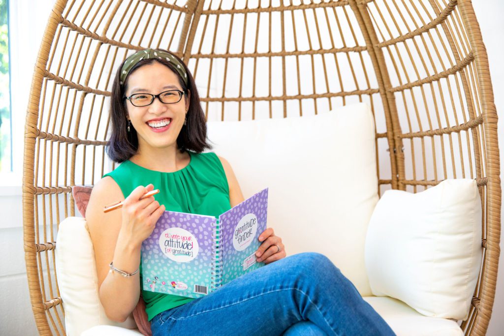 An Asian woman wearing a green top sitting in an egg chair smiling with her gratitude journal.