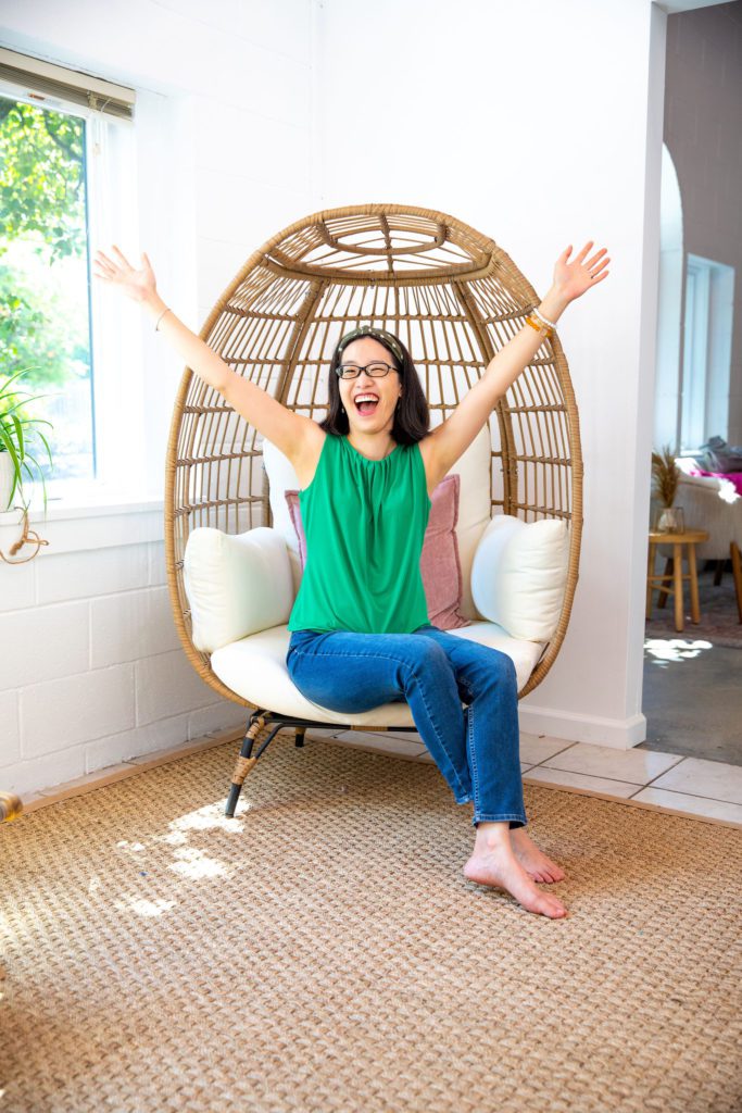 An Asian woman wearing a green top sitting in an egg chair with her arms up feeling excited.