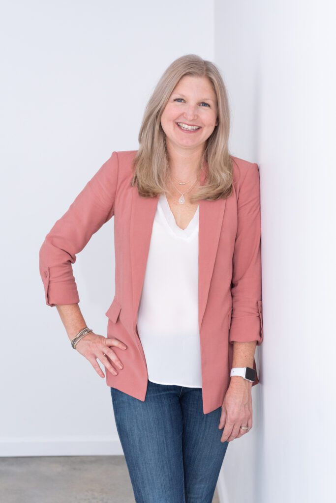 A female business coach smiling and leaning against a white wall with her hand on her hip.