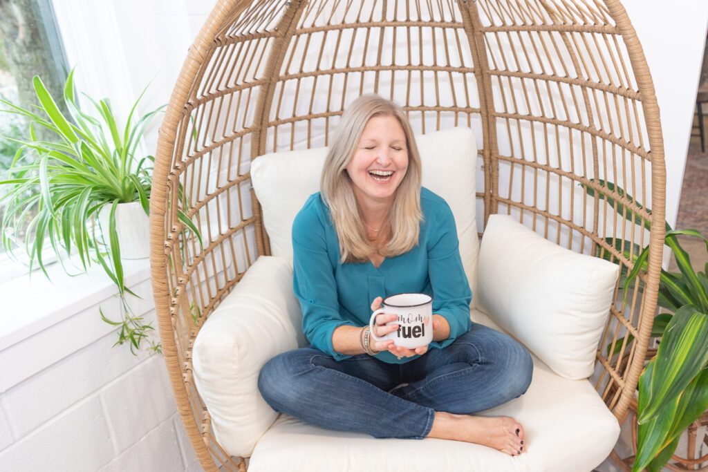 A female business coach sitting in an egg chair, laughing and holding a mug with "Mom Fuel" text.