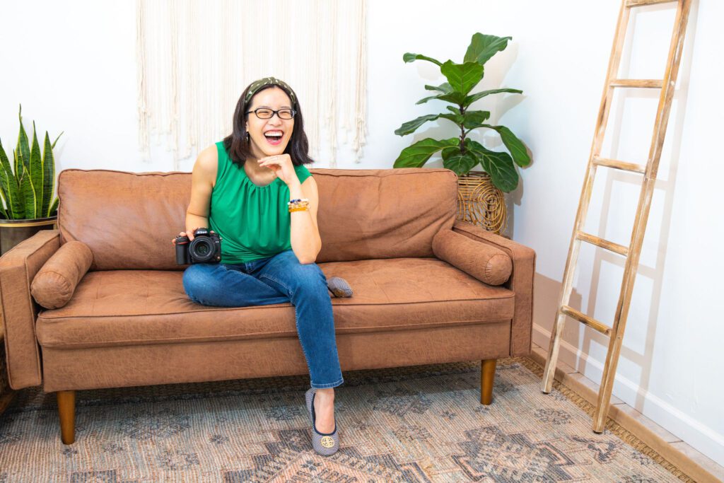 An Asian female personal branding photographer wearing a green top sitting on the floor and leading against a sofa with her DSLR camera.