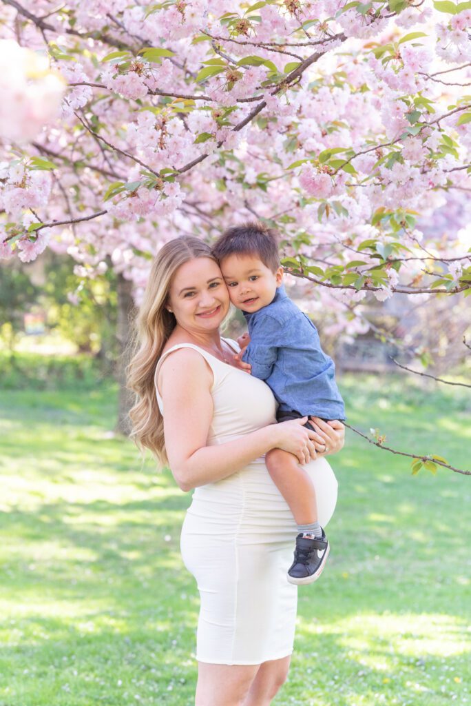 A pregnant mom is holding her toddler son under the cherry blossom tree.