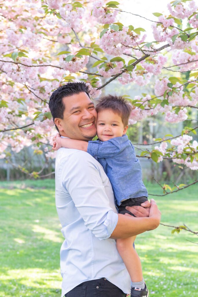 A dad is holding his toddler son under the cherry blossom tree.