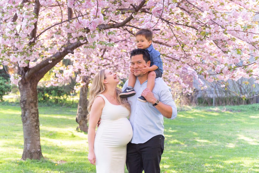 A family of 3 is smiling at each other while the toddler son sitting on the dad's shoulder under the cherry blossom trees.