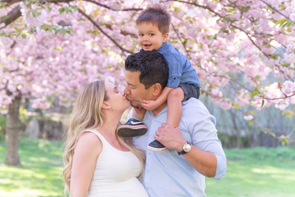 A woman and a man kissing with their toddler son sitting on the dad's shoulder under the cherry blossom trees.
