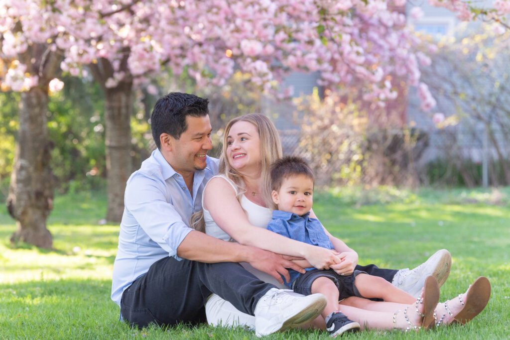 A family of three sitting on the grass and smiling for their maternity and family photoshoot under the cherry blossom tree.