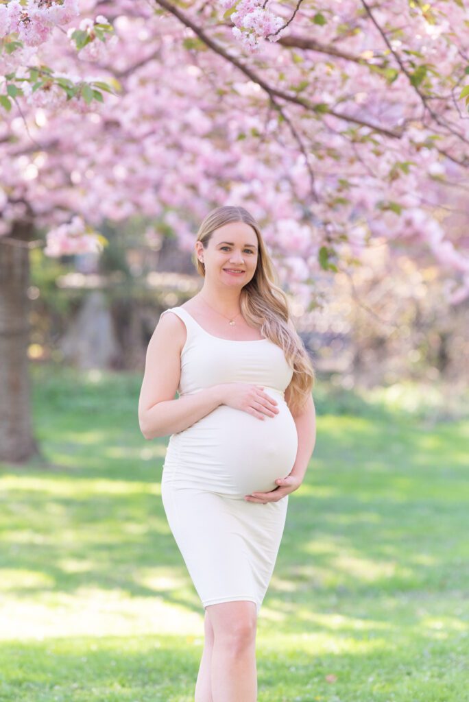 A pregnant woman in the off white dress holding her belly and posing for her maternity photoshoot under the cherry blossom tree.