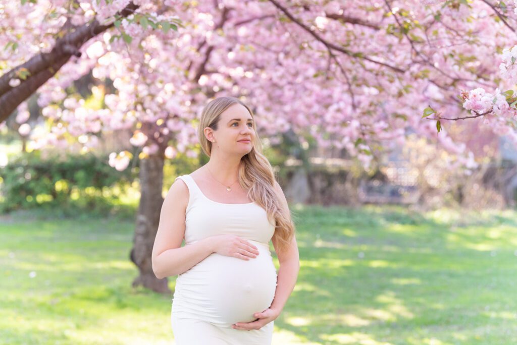 A pregnant woman in the off white dress holding her belly and posing for her maternity photoshoot under the cherry blossom tree.