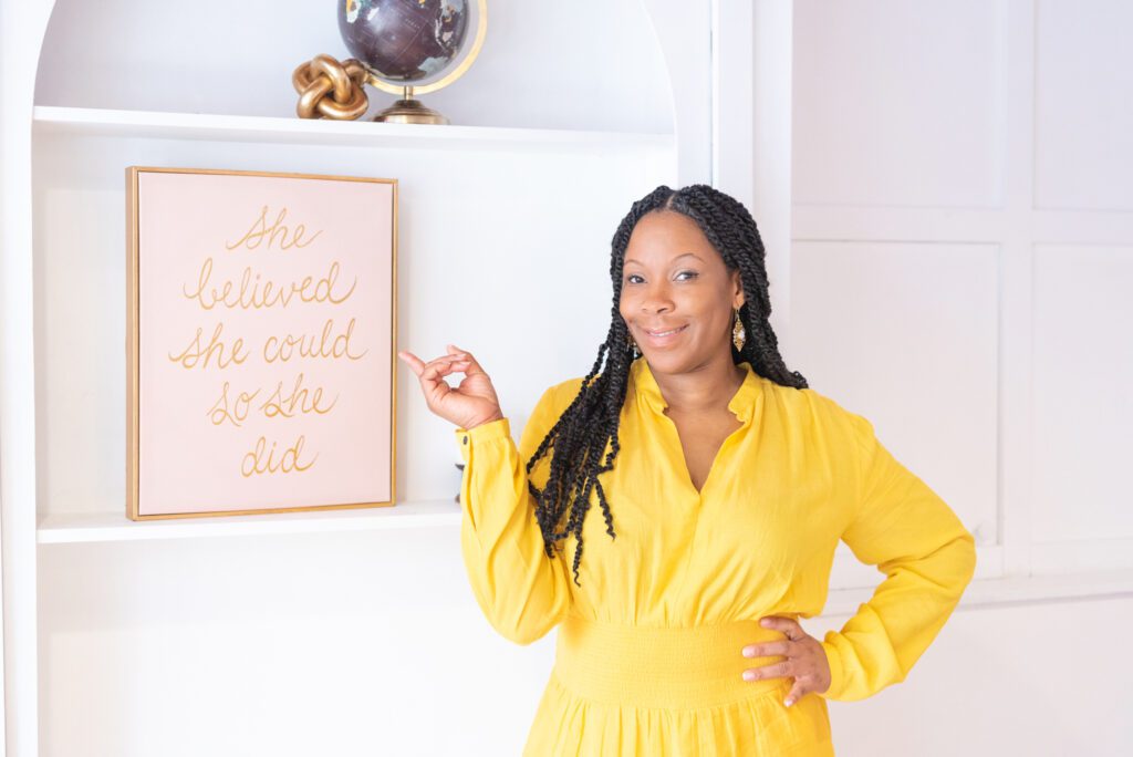 An African American woman in a yellow outfit pointing to a art piece with "She Believed She Could so She Did."
