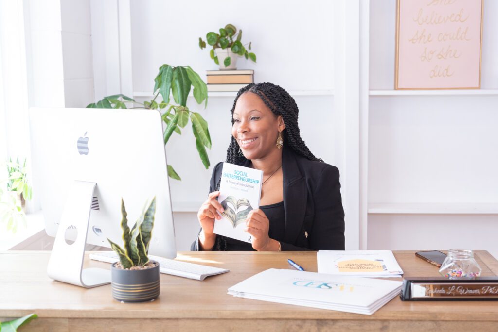 An African American woman business owner and author holding her book - Social Entrepreneurship: A Practical Introduction and showing someone on a computer.