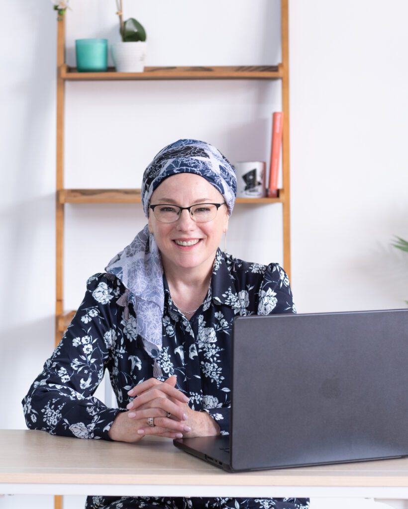 A Jewish woman sitting on a desk with her laptop and bookshelves in the background and smiling.