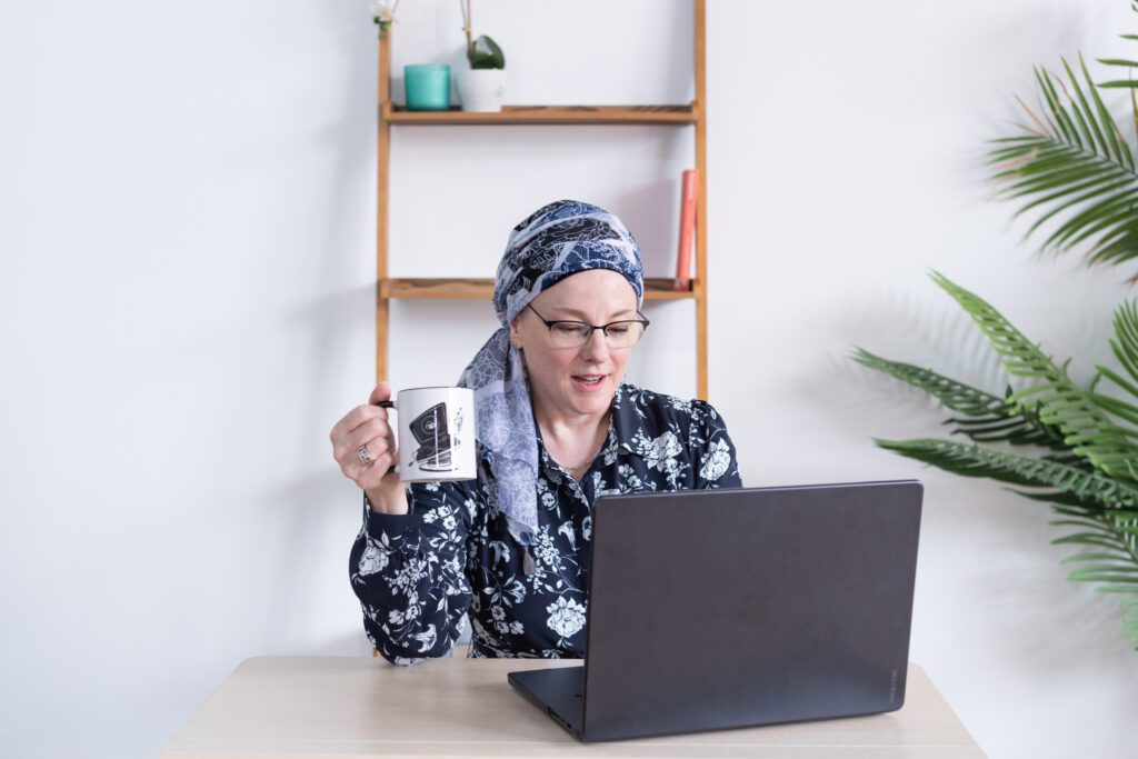 A Jewish woman sitting on a desk, holding a mug, and working on her laptop.