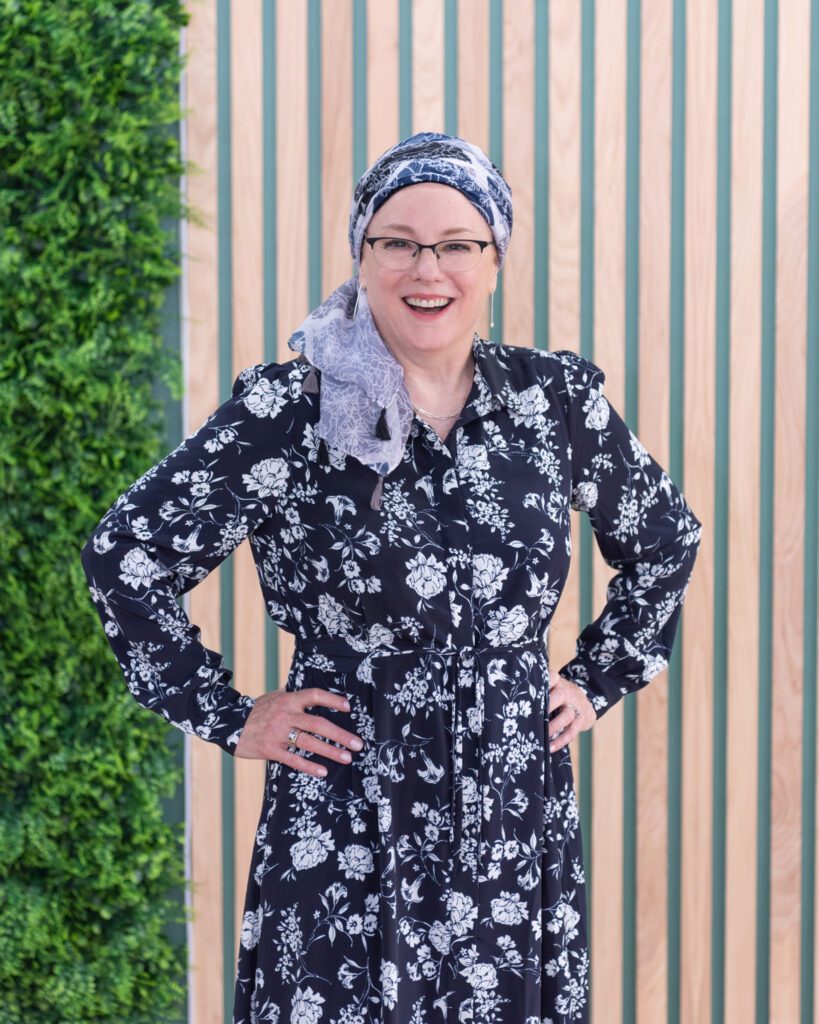 A Jewish woman standing in front of a fake plant backdrop with her hands on her hip and smiling.