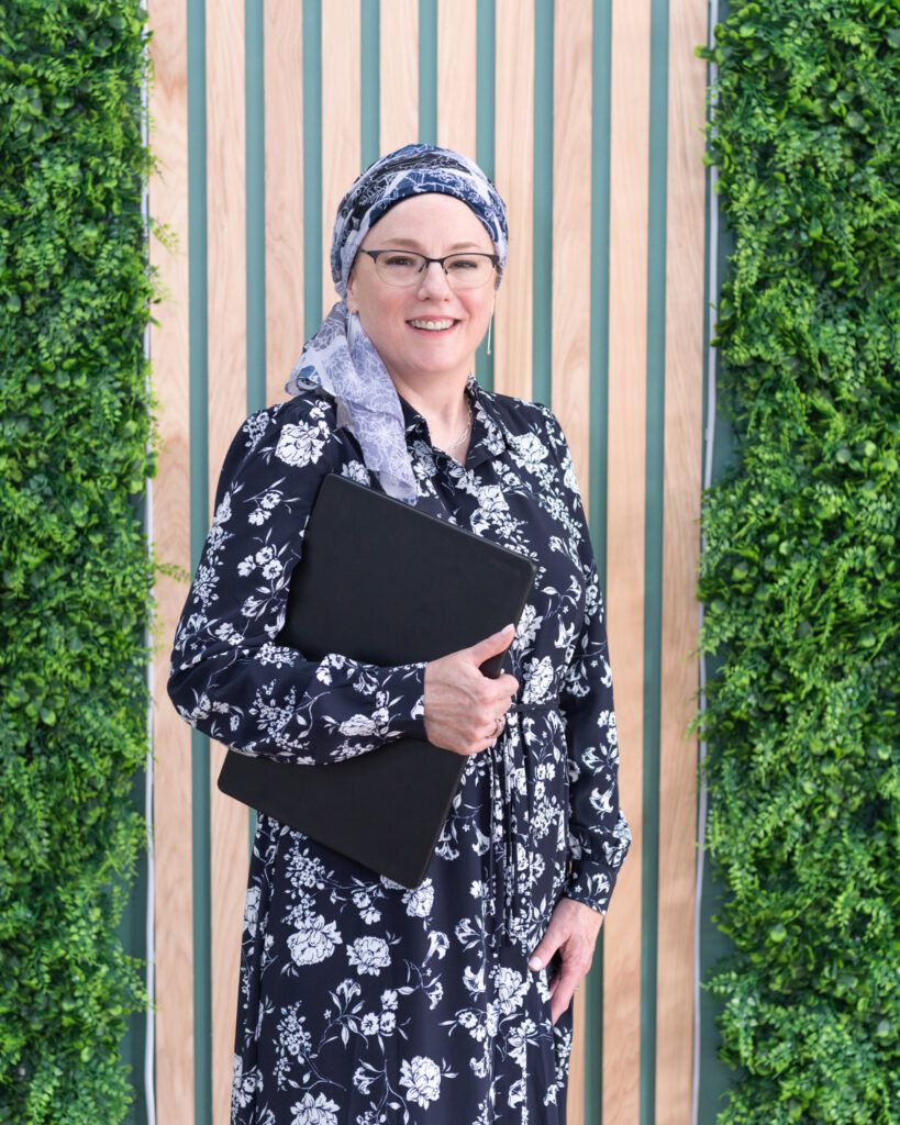 A Jewish woman standing in front of a fake plant backdrop holding her laptop.