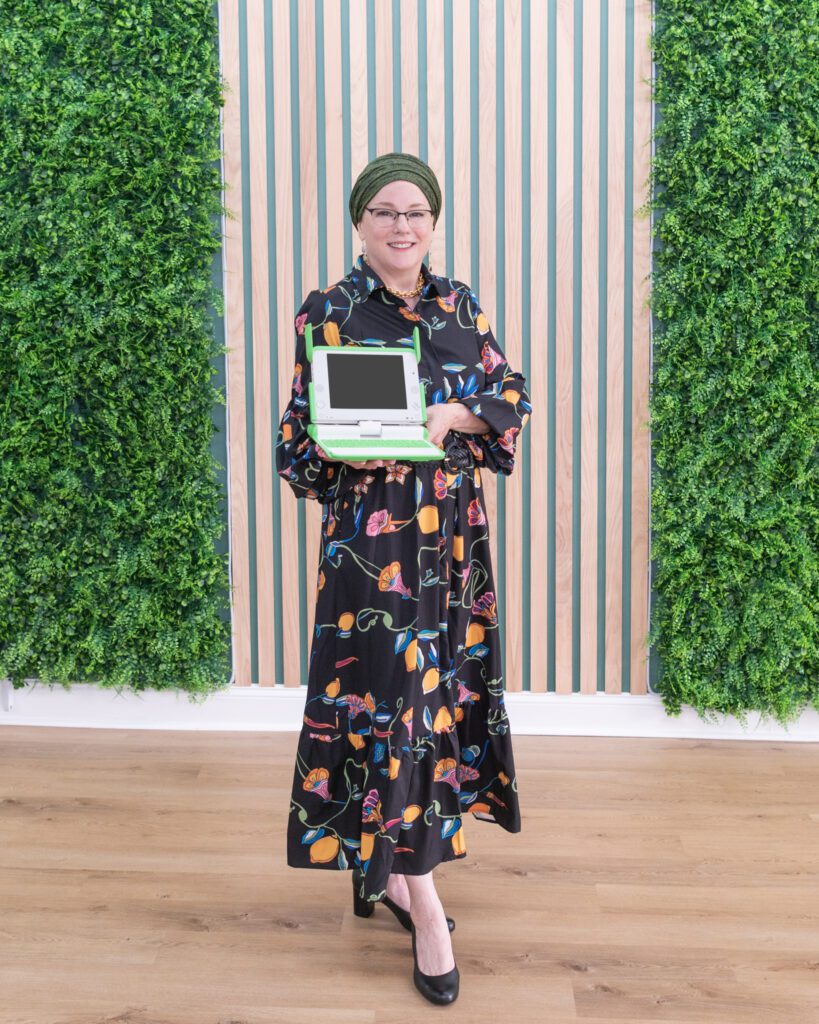 A Jewish woman standing in front of a fake plant backdrop holding a small programmable laptop.