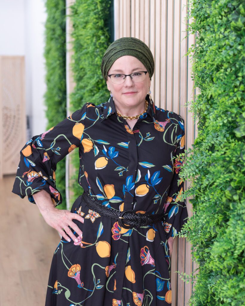 A Jewish woman leaning on a fake plant backdrop and smiling.