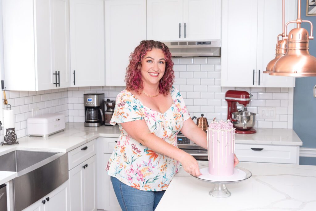 A female New Jersey cottage baker finish decorating a tall cake in her home kitchen.