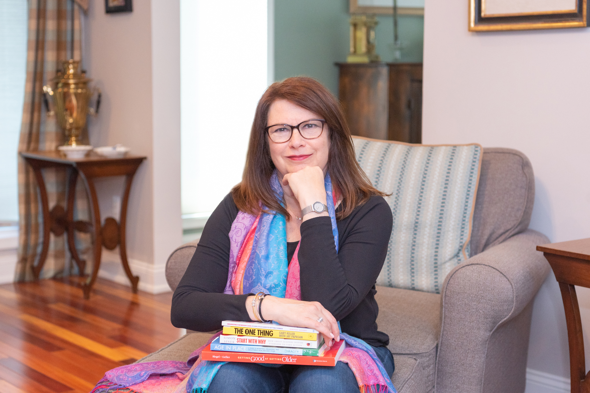 A female professional organizer sitting on a brown couch with a stack of books on her lap.